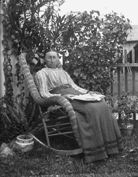 Mary Krueger is sitting outdoors near the side of a building in a rocking chair holding a book. Several potted plants are sittings on the ground beside her, and behind are vines and plants along a fence.