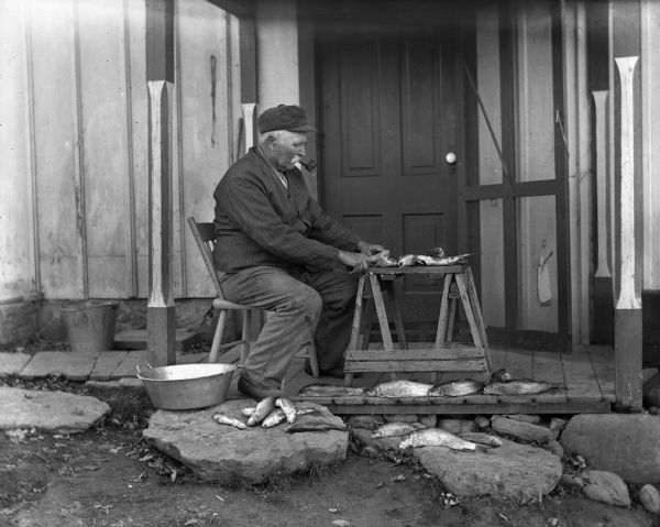 August Krueger sitting in a chair on the porch cleaning fish while smoking a pipe. Several of the fish are lying on the rocks next to the porch, and several others are lined up along the porch edge.