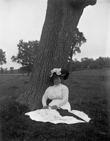 Sarah Krueger, sitting at the base of a tree, wearing an elaborate hat and a long white dress. She is holding an umbrella.