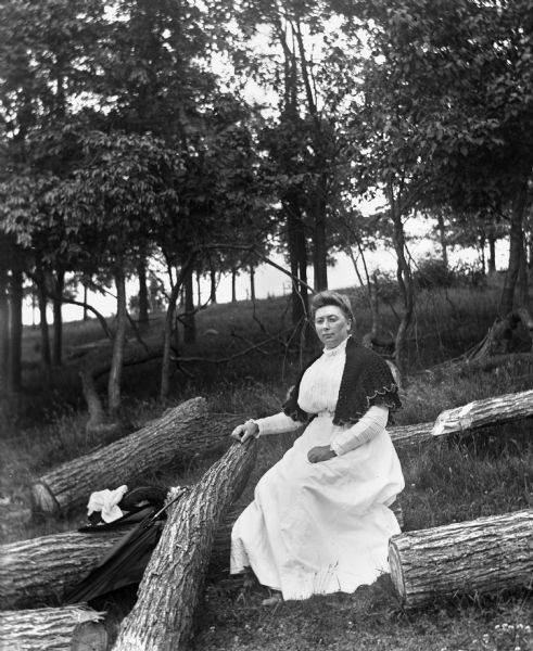 Outdoor portrait of Sarah Krueger sitting among several large tree logs on the side of a hill. Her hat and umbrella are resting on the logs next to her.