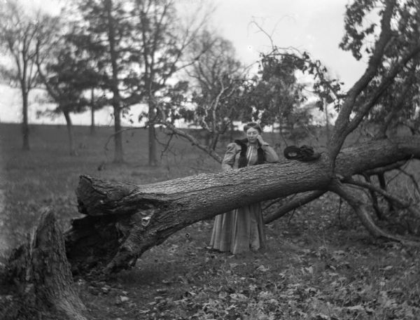 Sarah Krueger, standing and leaning her cheek on one elbow on top of a fallen tree. Her hat is sitting on top of the tree next to her.
