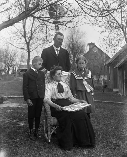 Outdoor group portrait of the Alexander Krueger family. Florentina Krueger is sitting in a wicker chair holding a photo album open on her lap. Alexander Krueger is standing behind her with his hand resting on the back of the chair, while Jennie and Edgar Krueger are standing on either side of Florentina. Farm buildings and a windmill are in the background.