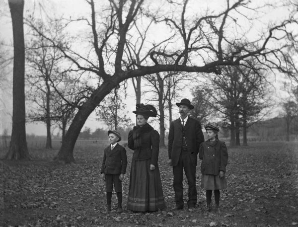 Outdoor group portrait of the Alexander Krueger family standing together in a wooded area. An oddly curved tree is behind the family. From left to right: Edgar, Florentina, Alexander, and Jennie Krueger.