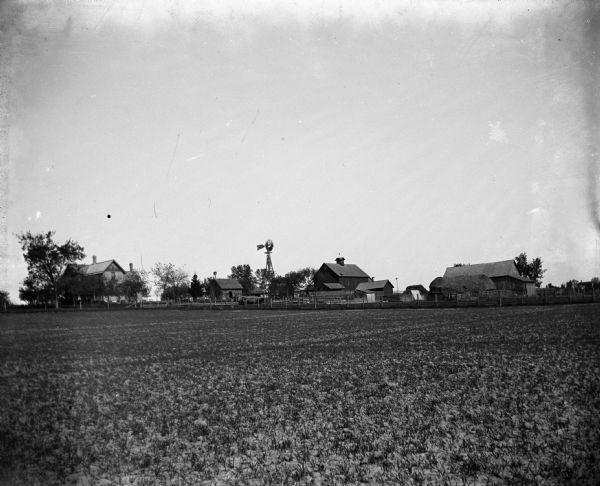 View across field from the south east of the Krueger farm, which includes farm buildings, fences, a farmhouse and a windmill.
