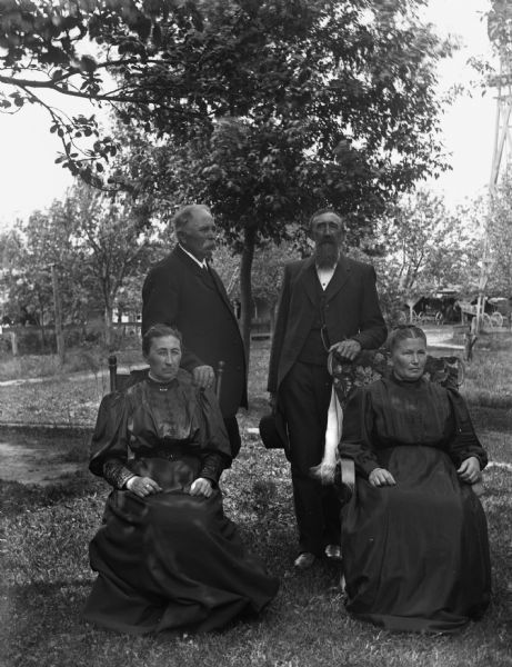 Outdoor group portrait of August and Mary Krueger and William and Bertha (Krueger) Goetsch. The two women are sitting in chairs, while their husbands are standing behind them resting their hands on the back of the chairs. In the far background horse-drawn carriages are near a building under trees.