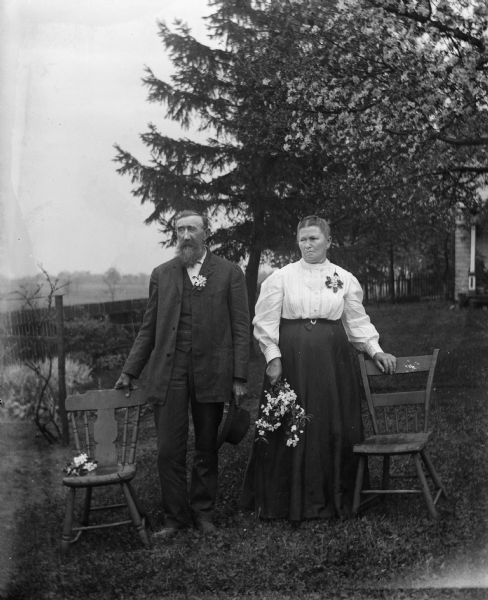 William Goetsch and his wife Bertha Krueger Goetsch standing outdoors between two chairs, each wearing a corsage. Bertha is holding a bouquet of flowers in her right hand. In the background is a fence, and the corner of a porch is on the right.