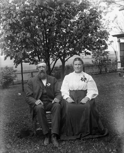 Outdoor portrait of William Goetsch and his wife, Bertha Krueger Goetsch, sitting in chairs on a lawn. They are each wearing a corsage, and William's hat is on the ground beside him. Behind them is a fence, and on the right, the corner of a porch.