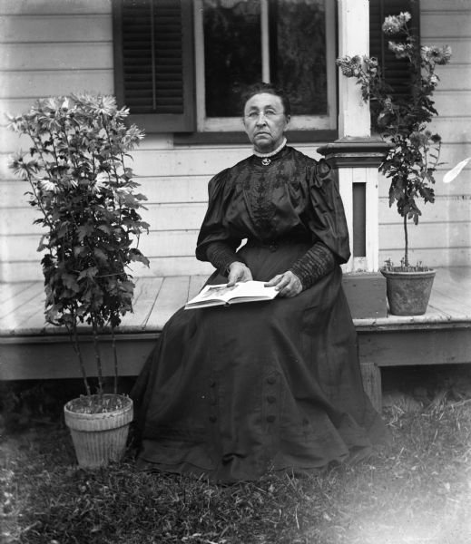 Mary Krueger sitting on the edge of the front porch with a book open in her lap. Potted plants are placed on either side of her.