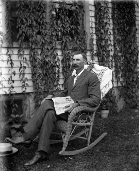 Portrait of Alexander Krueger sitting outdoors in the yard in a wicker chair with a newspaper open in his lap. His hat is on the ground next to him. The house behind him has windows covered in ivy.