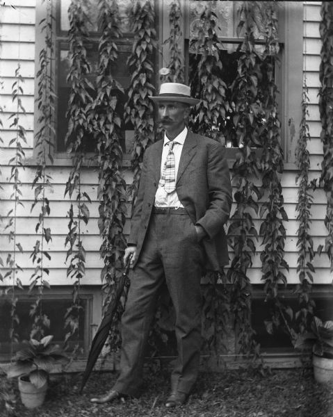 Outdoor portrait of Alexander Krueger standing in front of the ivy-covered windows of a house. He is wearing a hat and has his left hand in his pants pocket. In his right hand he is holding an umbrella like a cane. A potted plant is on the ground on the left.