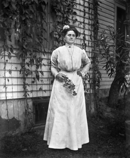 Outdoor portrait of Florentina Krueger holding a bouquet of flowers and standing at the side of a house with ivy hanging over the windows. She is wearing a long white dress and a flower in her hair.