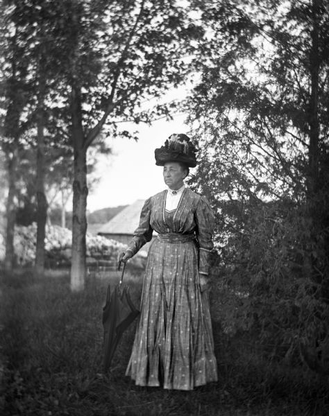 Outdoor portrait of Mary Krueger standing along a tree line, wearing an elaborate hat and holding an umbrella. Behind her is a farm building.