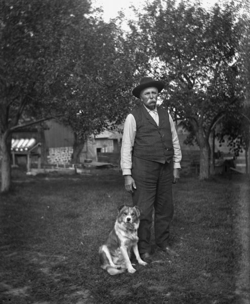 August Krueger standing outdoors in a yard, with a dog sitting at his feet. Trees and farm buildings are in the background.
