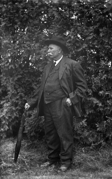August Krueger wearing a suit, hat, and holding an umbrella, standsingin profile in front of a large pine tree.