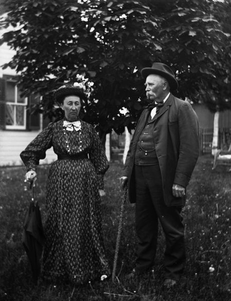 August and Mary Krueger standing together in the yard, each wearing hats. Mary is holding and supporting herself with an umbrella in her right hand, and August is holding a cane in his right hand.