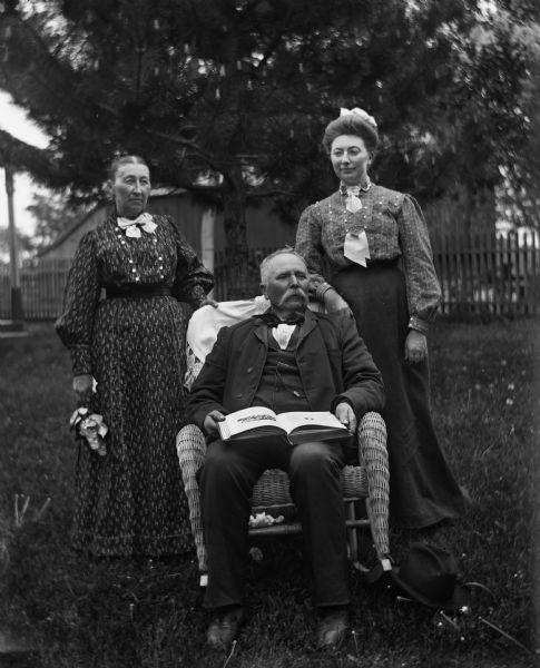 Outdoor portrait of August Krueger sitting in a wicker rocking chair supporting an open book on his lap, while Mary and Sarah Krueger are standing behind him. A fence and farm buildings are in the background.
