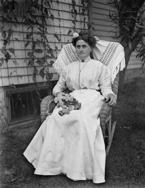 Outdoor portrait of Florentina Krueger sitting in a wicker rocking chair in the yard near the side of an ivy-covered house. She is wearing a long white dress and has a flower in her hair. There is a bouquet of flowers draped across her lap.