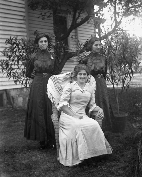 Group portrait of Hattie Fels, sitting in a wicker rocking chair, with Josie and Esther Bigalk standing behind her. Two potted plants are on the lawn, and in the background is the side of a house.