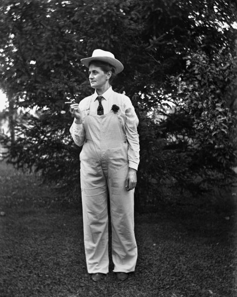 Florentina Krueger posing outdoors in white overalls, tie, and mens hat with a cigar propped in her left hand.