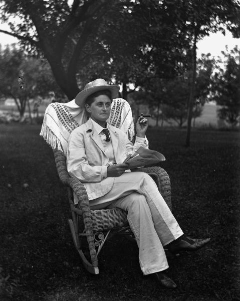 Portrait of Florentina Krueger sitting outdoors in a wicker chair. She is wearing a man's white suit and hat, and is holding a cigar in her left hand and a newspaper in her right hand.