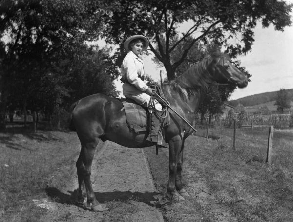 Flora Fels Weege sitting on top of a horse holding a rifle. She is wearing a men's suit, a hat, and a belt holding ammunition.