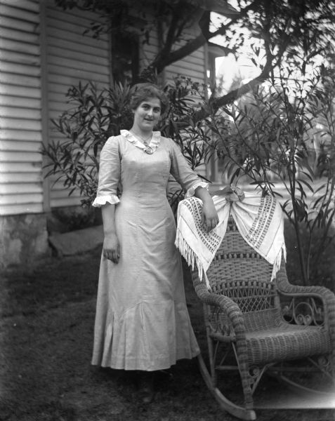 Hattie Fels Owen standing outdoors next to a wicker rocking chair. Several potted plants are placed behind her, and the side of a house is in the background.