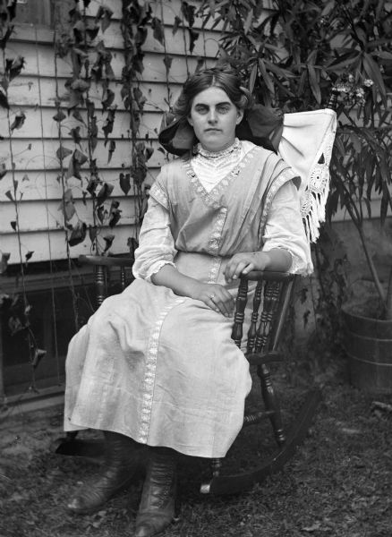 Outdoor portrait of Flora Fels Weege sitting in a rocking chair next to the Krueger house. A large potted plant is on the ground behind her.