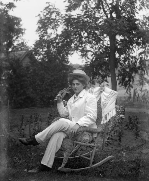 Hattie Fels Owen sitting in a wicker rocking chair. She is wearing a man's suit and hat and posing with a cigar.