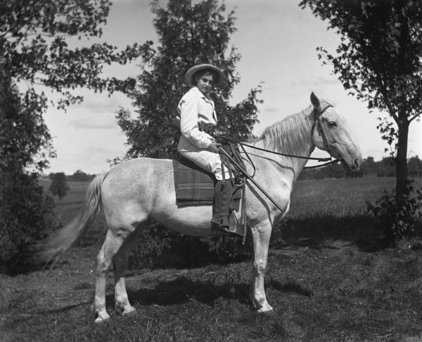 Flora Fels Weege sitting on a white horse while wearing a man's suit, hat, and a belt containing ammunition. She has a rifle propped in her right arm.
