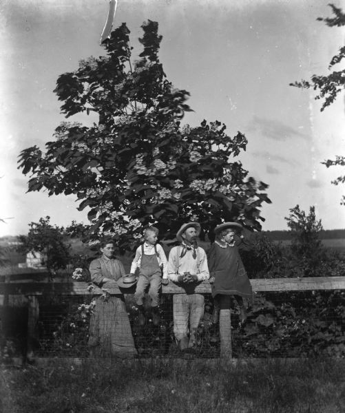 Outdoor portrait of the Alexander Krueger family posing along a fence in front of a Catalpa tree. Jennie and Edgar Krueger are sitting on top of the fence while Florentina and Alexander are leaning against it.