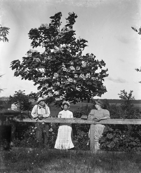 Outdoor portrait of the August Krueger family posing with farming tools and leaning on a fence in front of a Catalpa tree. From the left: August Krueger is smoking a pipe and standing next to a pitch fork, Mary Krueger is holding a rake, and Sarah Krueger is standing next to a hoe.
