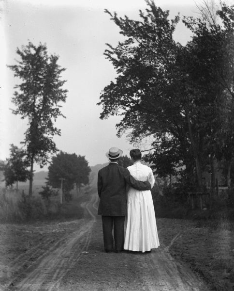 Rear view of Florentina Krueger strolling down a road with Tillie Timmel. Florentina is dressed in a man's suit and has her arm around Tillie's waist.