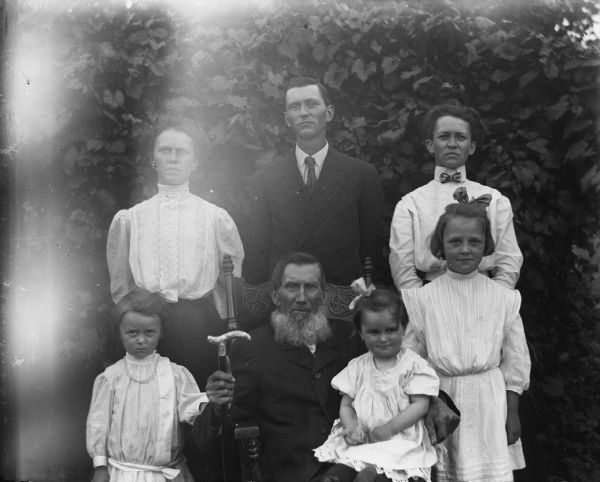 Outdoor portrait of the William Wendorf family. William is sitting in a rocking chair holding a cane with Mabel Kyzowski in his lap, while Ruth and Alice Kressin are standing on either side of him. Behind them are Martha Wendorf Kressin, Ed Wendorf, and Minnie Wendorf Kyzowski.