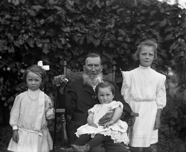 Outdoor portrait of William Wendorf with his granddaughters. William is sitting in the middle on a rocking chair holding a cane. Mabel Kyzowski is sitting in his lap, while Ruth and Alice Kressin are standing on either side of him.