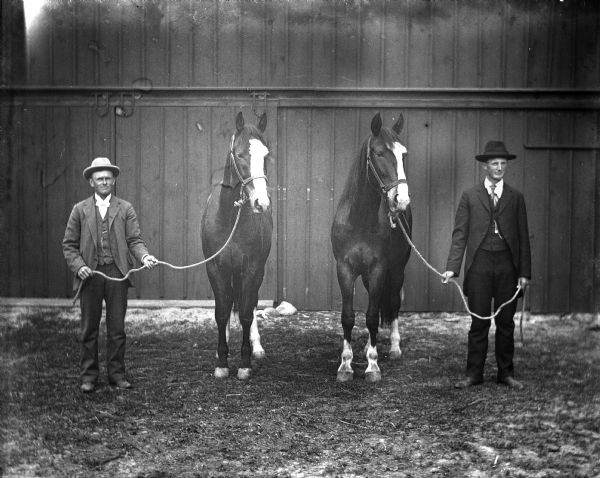 Gust Wendorf and William Will standing in front of a barn with their horses. Each of the men are holding the rope attached to the horse's halter.