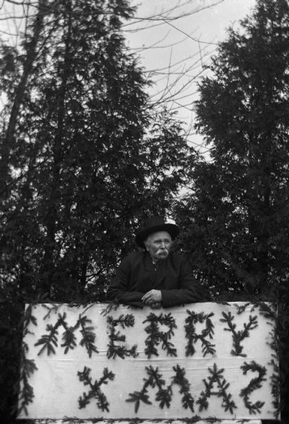 August Krueger standing behind a Merry Xmas sign made from pine branches.