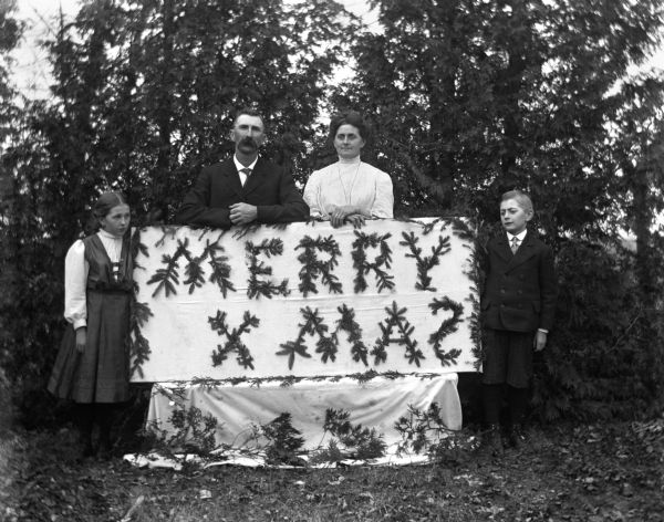 Outdoor group portrait of the Alexander Krueger family with a Merry Xmas sign made from pine branches. Alexander and Florentina are standing behind the sign while the twins, Jennie and Edgar, are standing on either side.