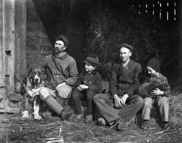 Alexander Krueger, Edgar Krueger, Frank Goetsch, and Anson Goetsch sitting together on the edge of the open hay barn door. Alexander has his arm wrapped around a large dog, while Anson is holding a grey cat in his lap.