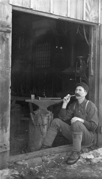 Alexander Krueger sitting in the open doorway to a blacksmith shop smoking a pipe.