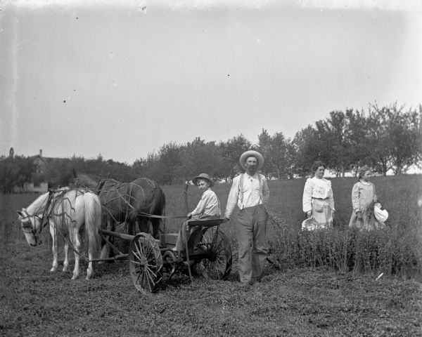 Alexander, Edgar, Florentina, and Jennie Krueger cutting alfalfa using a horse-drawn mower. Edgar is sitting on the mower holding the reins of the two-horse team, while Alexander is standing behind him leaning on the mower and holding some alfalfa. The two women are standing in the uncut field. In the background on the right are farm buildings.