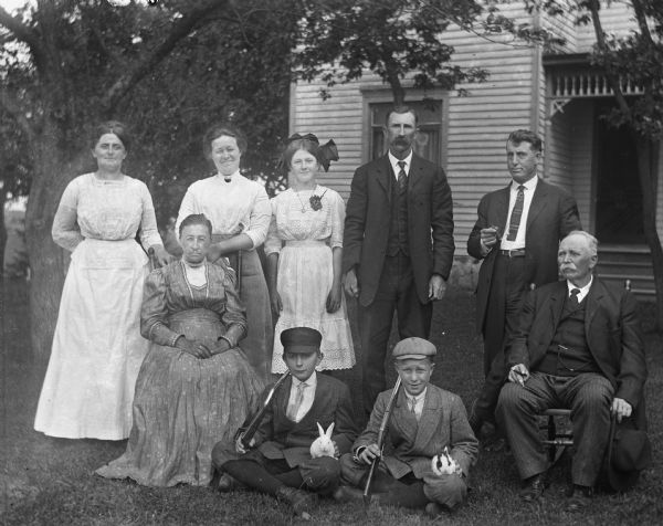 Outdoor group portrait of Florentina, Jennie, Alexander, August, Mary, and Edgar Krueger with Ed Buening, Mrs. Buening, and their son in front of the Krueger household. The two young boys are sitting up front holding small rabbits in their laps and rifles propped over their right shoulder.