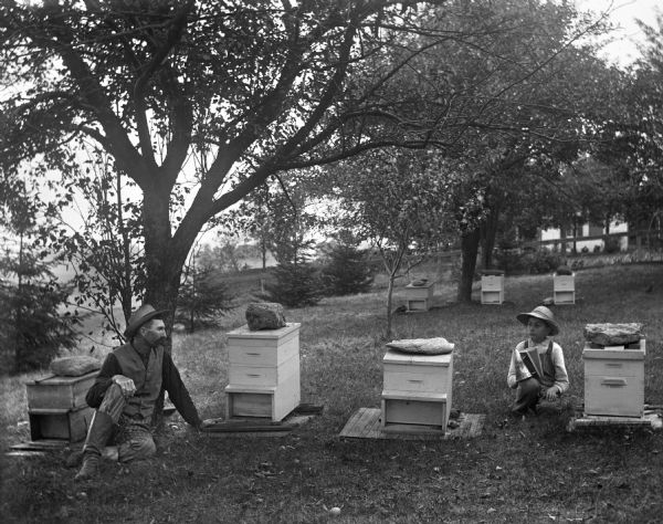 Alexander and Edgar Krueger sitting among a group of beehives. Edgar is holding a smoker in his hand. Behind them in the background is a building.