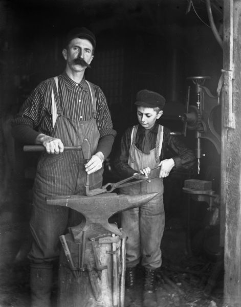 Alexander and Edgar Krueger posing together as blacksmiths in a blacksmith shop. Edgar is steadying the piece of metal on the anvil while Alexander is using a hammer and chisel on the metal piece.