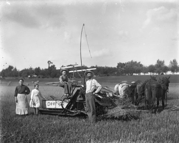 The Alexander Krueger family posing with a Deering Binder being pulled by a three-horse team through a field. Florentina, Jennie, and Alexander are standing behind the binder while Edgar is sitting in the drivers seat.