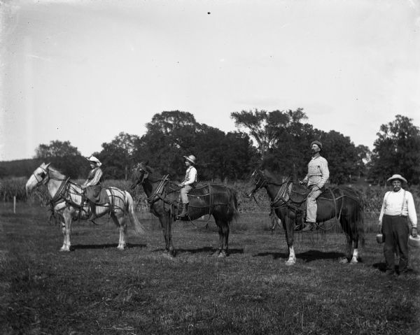 Jennie, Edgar, and Alexander Krueger posing bareback on horses. They are lined up to face the left, spaced in a row. The horses are wearing horse collars, blinders and fly-nets. August Krueger is standing at the end of the line on the right holding what may be rolls of twine in each hand.