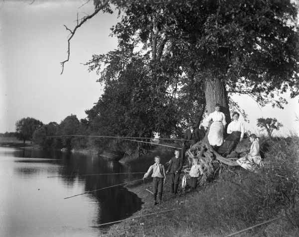Several members of the Krueger and Backhause families fishing along the bank of the Rock River. Mr. and Mrs. Backhouse and Florentina and Jennie Krueger are sitting along the roots of a large tree while Edgar Krueger and the two Backhause boys are standing along the edge of the water with their fishing poles.