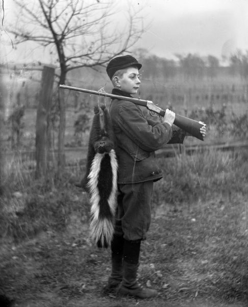 Edgar Krueger posing with a skunk he caught. The skunk is hanging from the rifle he has resting on his shoulder.