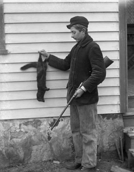 Edgar Krueger posing with a mink he trapped. He is looking down at the mink, which he is holding up against the house, while holding his rifle in the other hand.