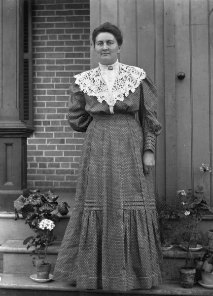 Outdoor portrait of Ida Scholz standing in front of the Langholf home. Several potted plants are on the steps beside her.