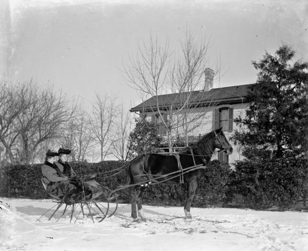 Ella and Ida Scholz sitting in a cutter (sleigh) in front of the Langholf home. The sleigh is being pulled by a horse.